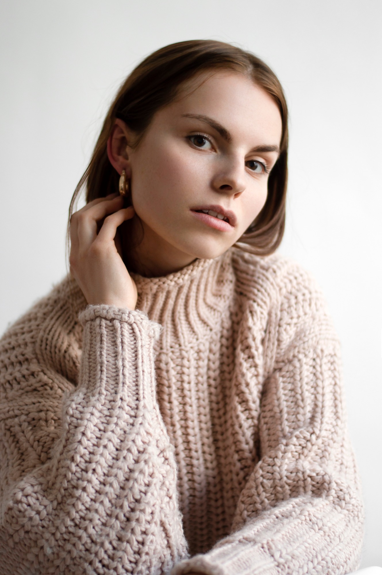 Portret kobiety w beżowym, grubym swetrze, z dłonią przy uchu ozdobionym złotym kolczykiem, na jasnym tle.