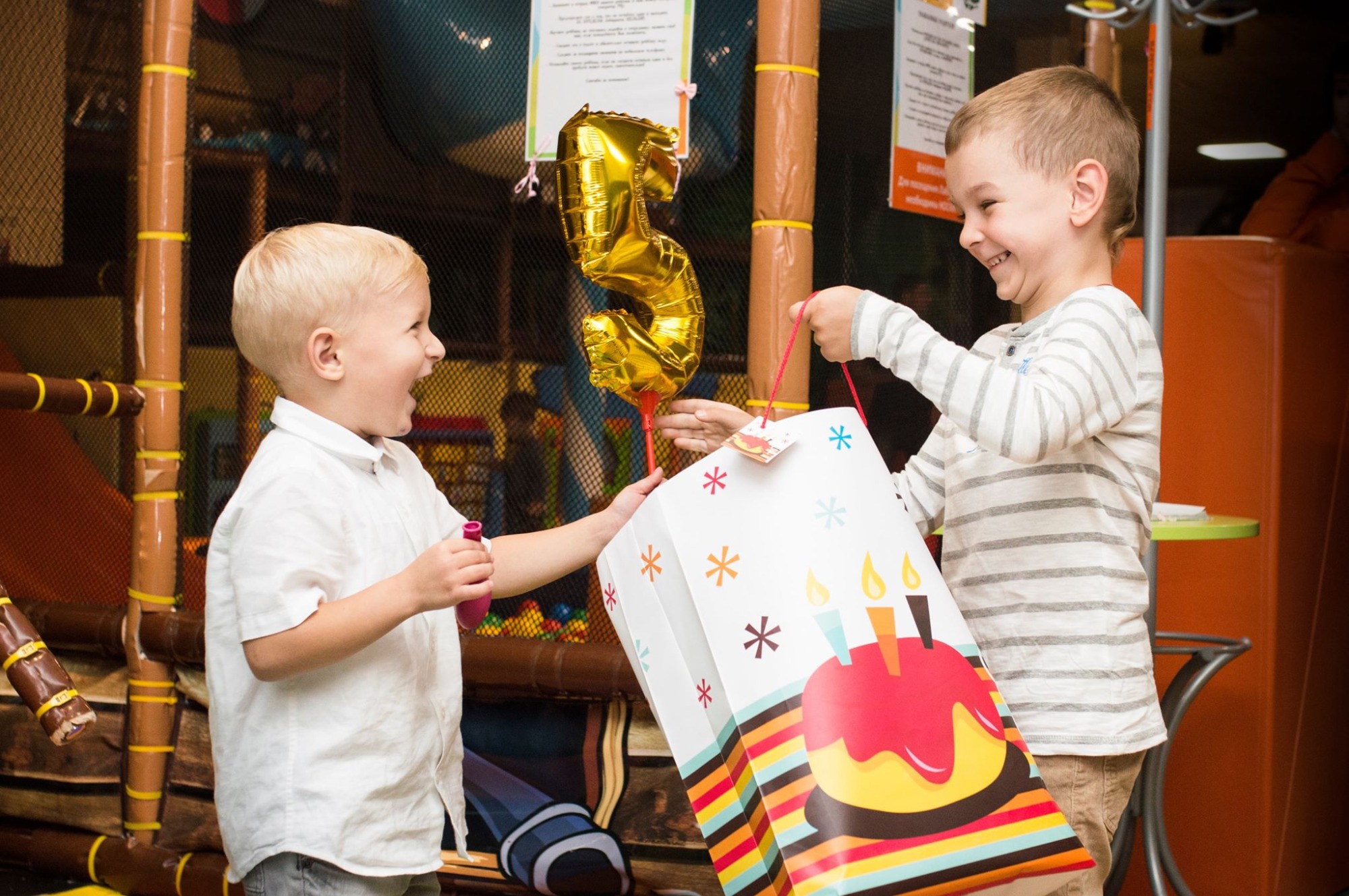 Dwóch uśmiechniętych chłopców w sali zabaw, jeden trzyma balon w kształcie cyfry pięć, drugi podaje mu torbę prezentową z motywem tortu.