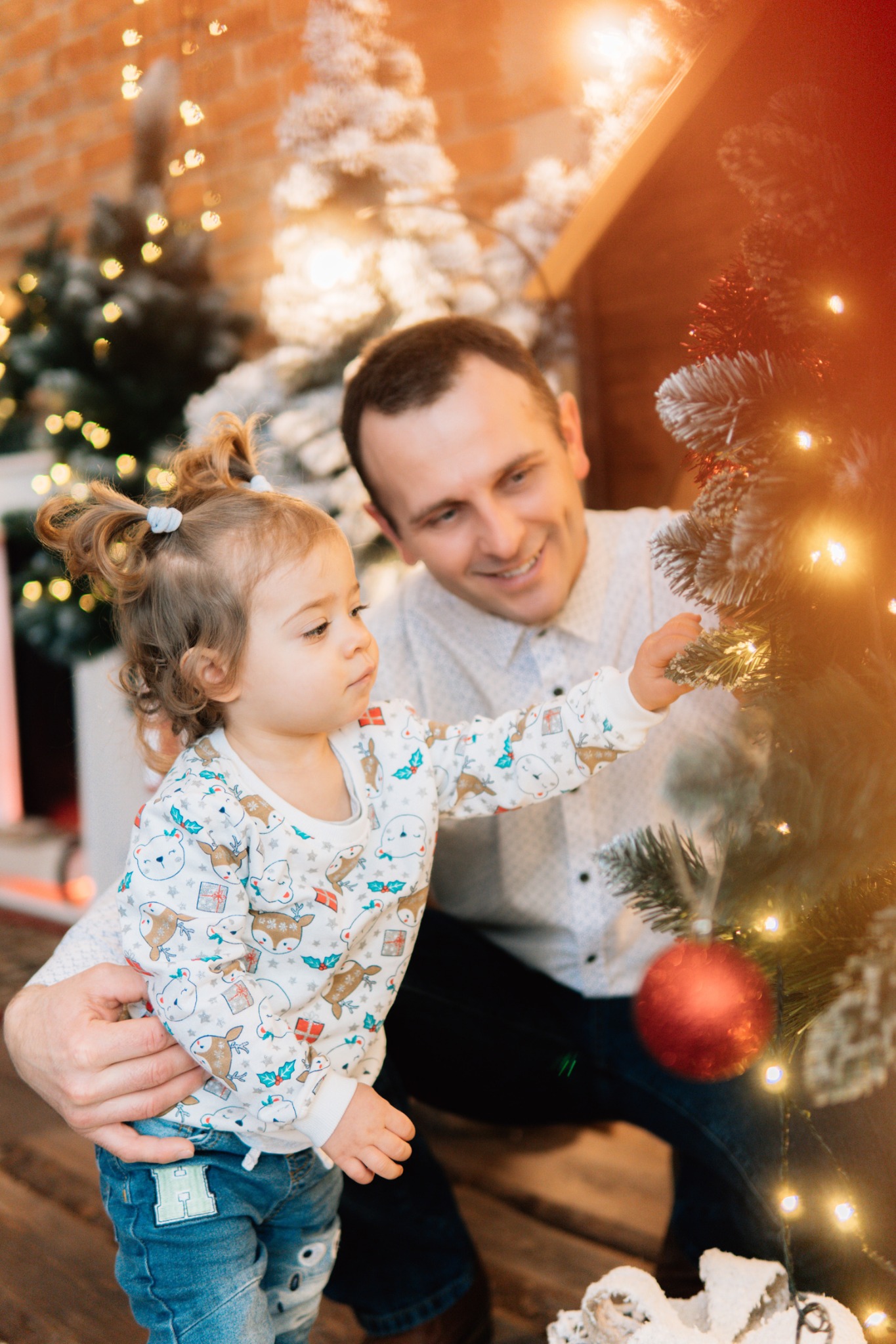 Tata z córeczką dekorują świąteczne drzewko, dziewczynka w bluzie z motywem reniferów dotyka gałązki, w tle ceglana ściana i migoczące światełka