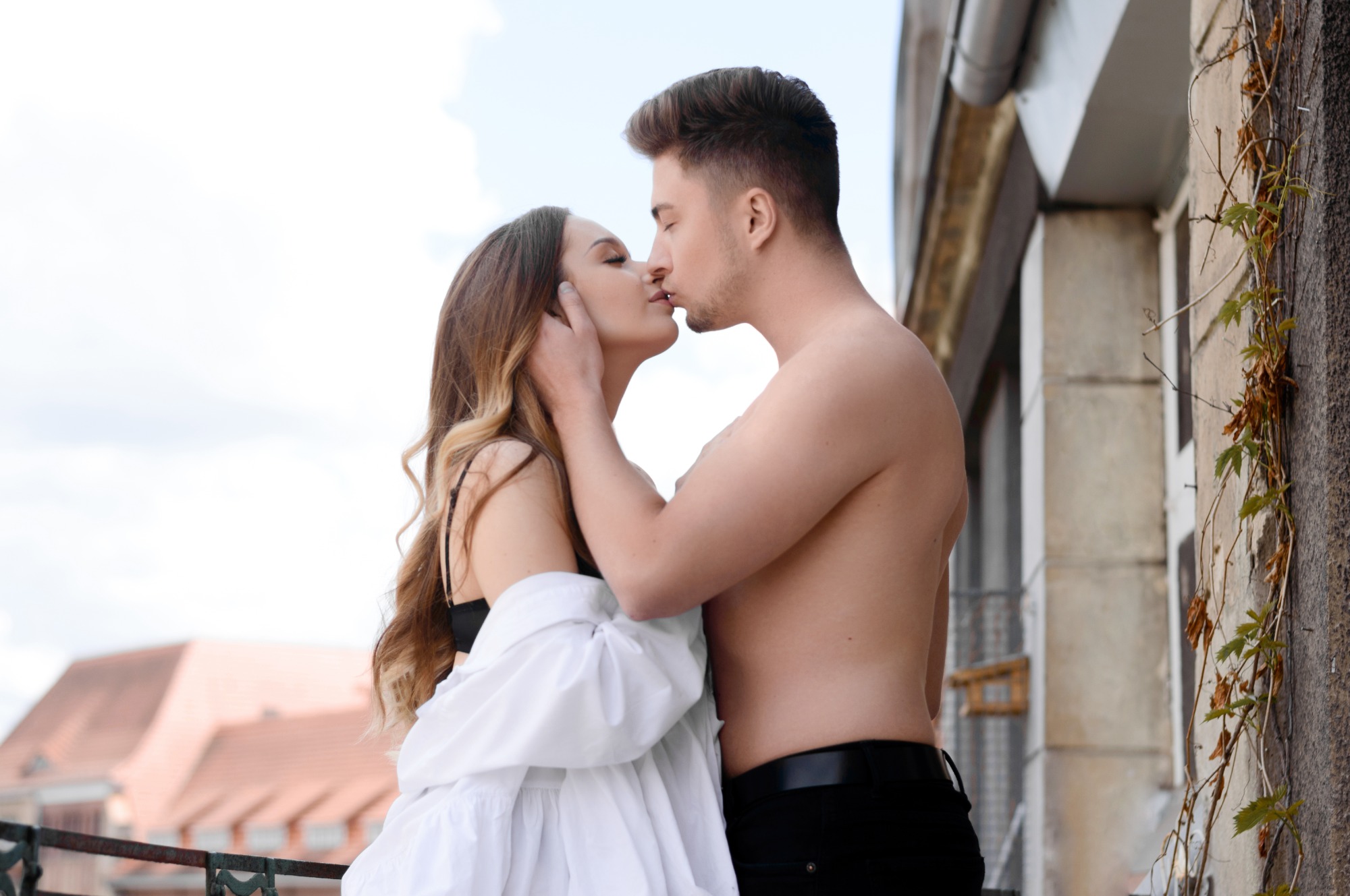 Para całująca się na balkonie z widokiem na dachy budynków; kobieta w białej koszuli i czarnym staniku, mężczyzna bez koszuli, oparcie balkonu z metalowych prętów, zarośnięta bluszczem ściana budynku.