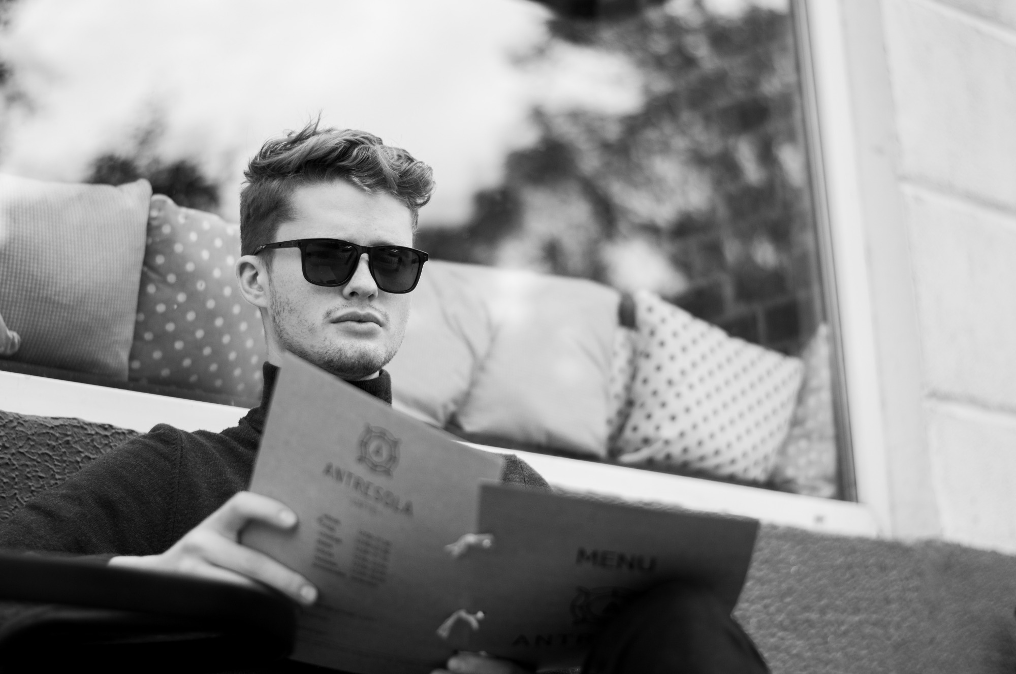 Czarno-białe zdjęcie mężczyzny w okularach przeciwsłonecznych siedzącego na zewnątrz i przeglądającego menu restauracji 'Antresola', z poduszkami w tle.