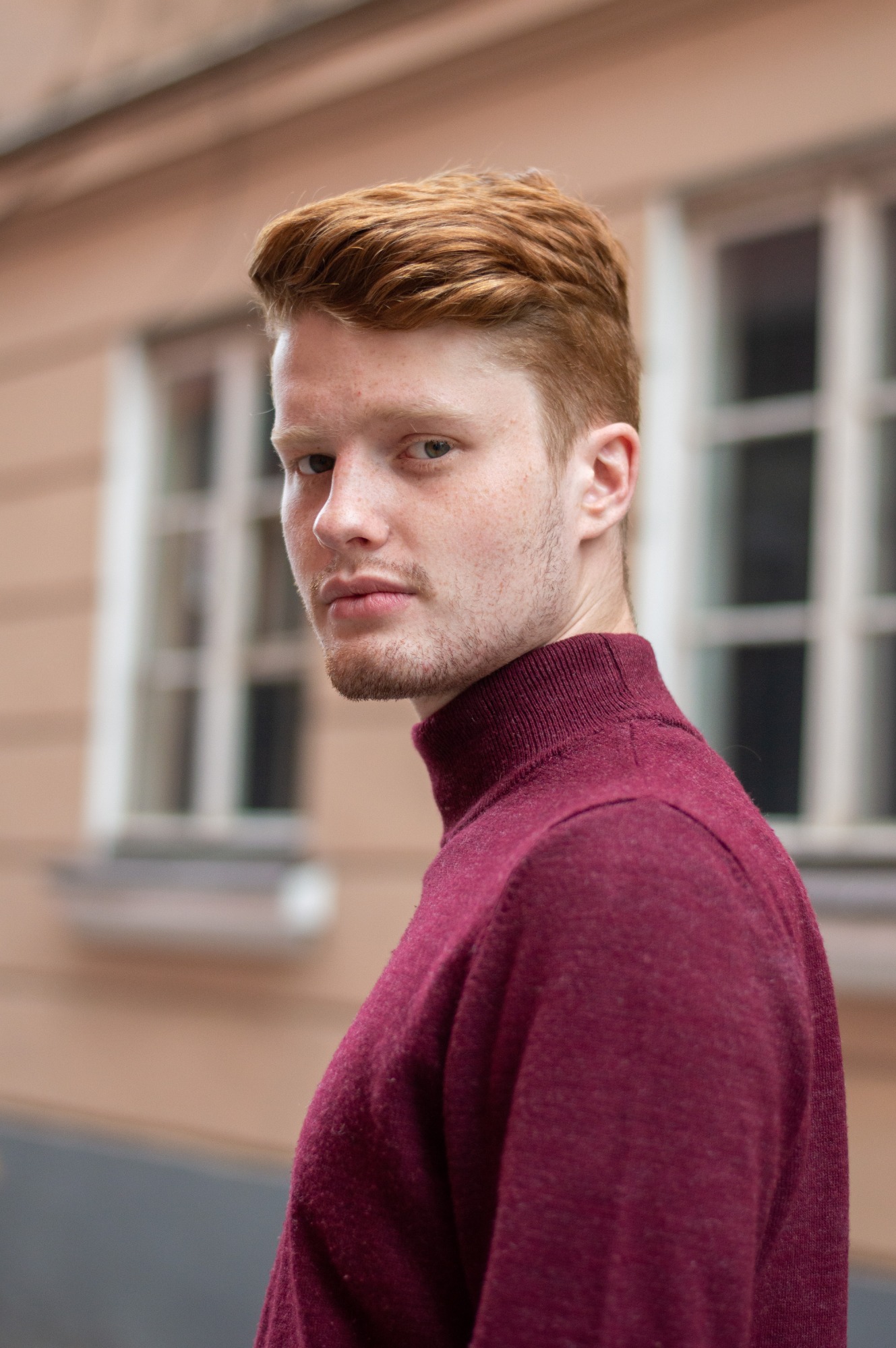 Portret rudowłosego mężczyzny w bordowym golfie, odwracającego się i spoglądającego w obiektyw na tle budynku z oknami.
