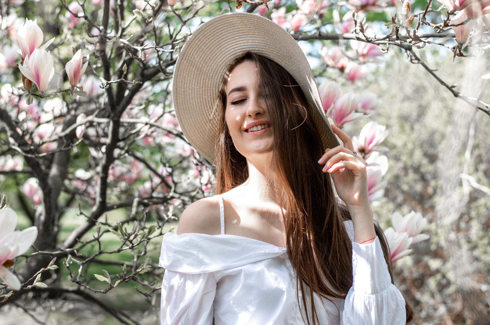 Uśmiechnięta brunetka w białej bluzce i słomkowym kapeluszu na tle kwitnącej magnolii w słoneczny dzień.
