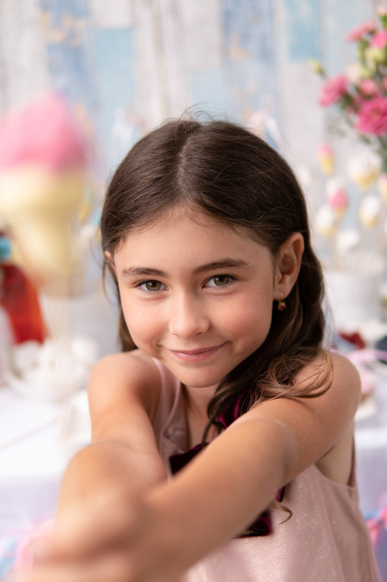 Portret uśmiechniętej dziewczynki z brązowymi włosami i zielonymi oczami, na tle dekoracji z waty cukrowej i kwiatów.