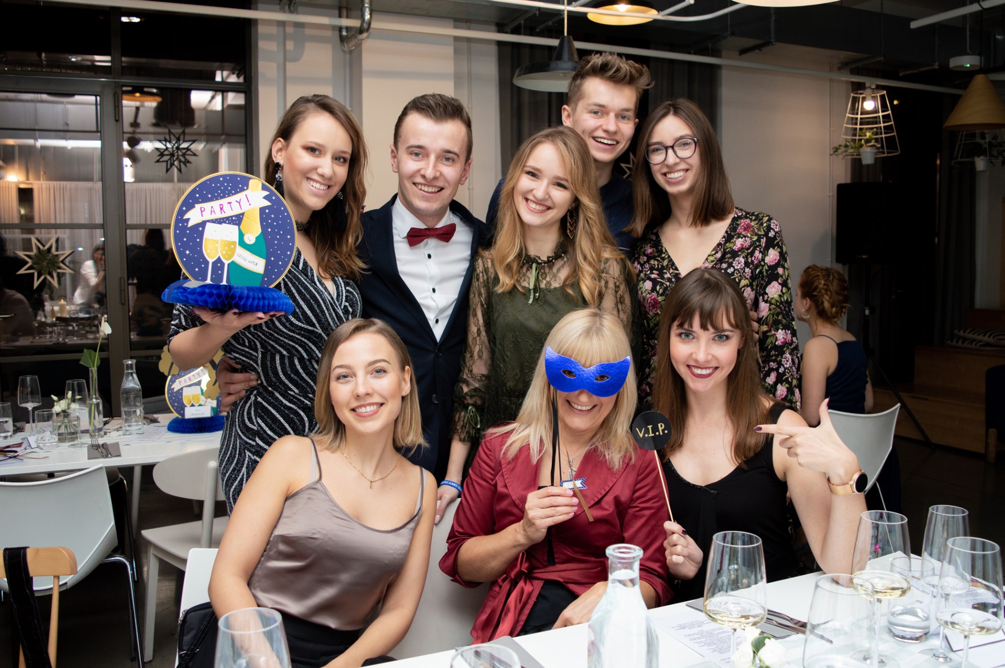 Grupa uśmiechniętych osób pozuje do zdjęcia podczas imprezy, z dekoracjami i rekwizytami, w tle widoczne nakryte stoły i okno.