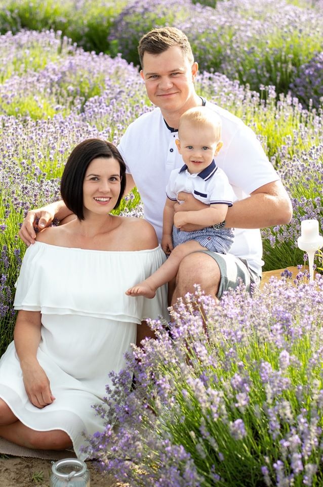 Uśmiechnięta rodzina - mama w białej sukience, tata i syn w białych koszulkach - pozuje na tle pola lawendy z piknikowym koszem i świecą.