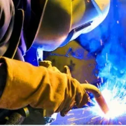 Spawacz w ochronnym hełmie i rękawicach spawalniczych podczas pracy, z intensywnym niebieskim dymem i iskrami