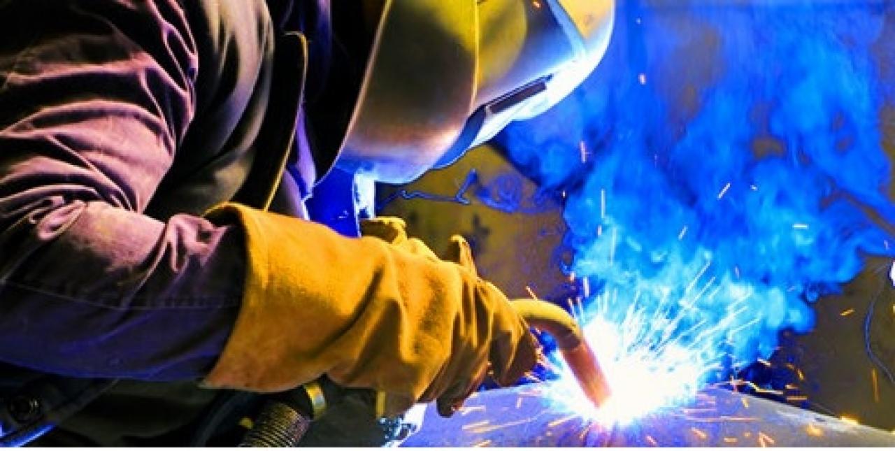 Spawacz w ochronnym hełmie i rękawicach spawalniczych podczas pracy, z intensywnym niebieskim dymem i iskrami