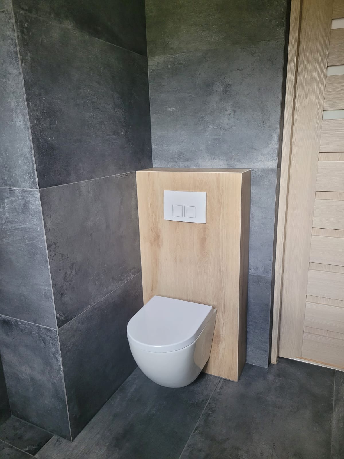 Nowoczesna toaleta podwieszana z drewnianą obudową i szarymi płytkami w łazience. Minimalistyczny design, widoczne płytki i fragment drzwi.