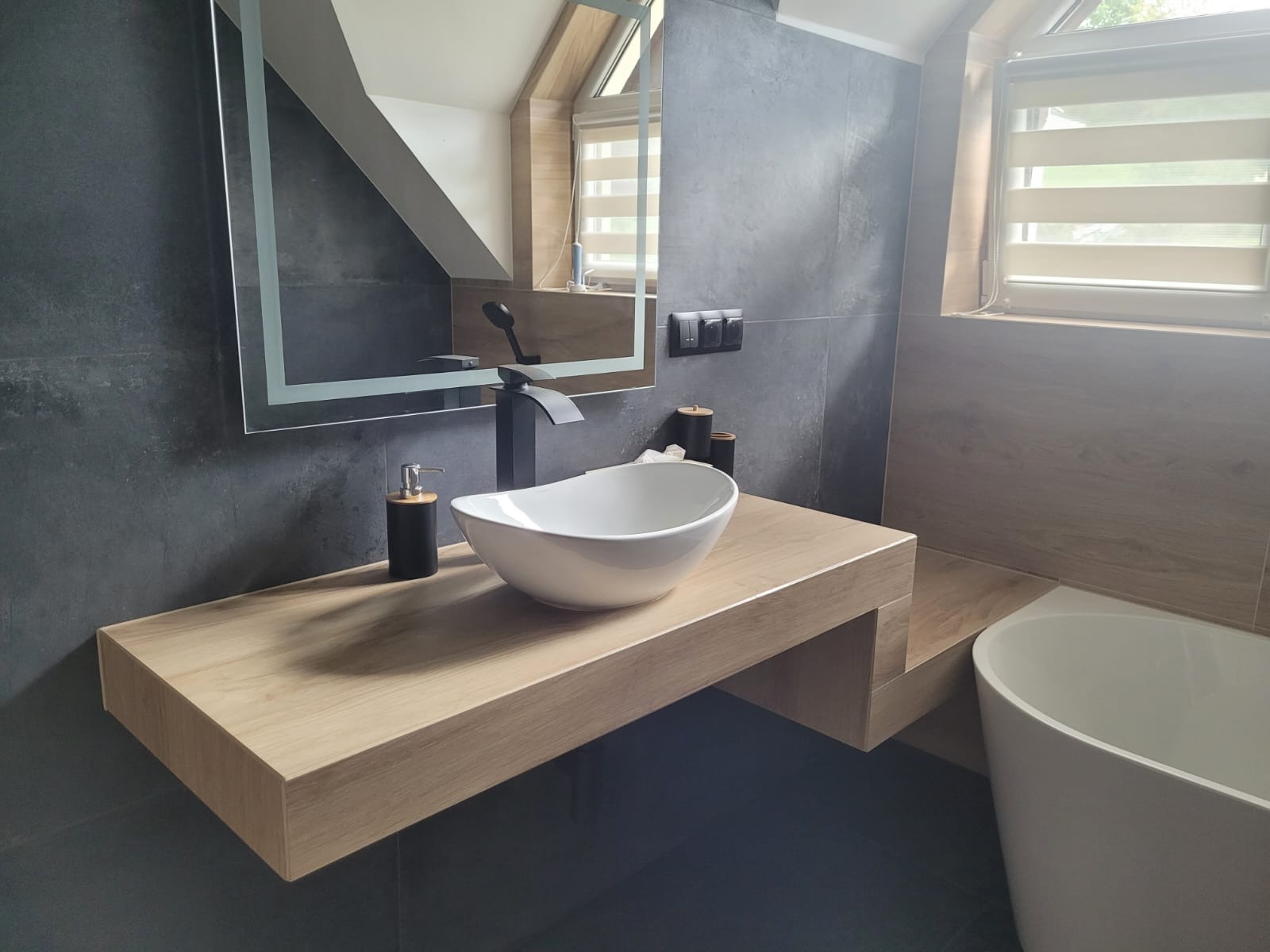 Nowoczesna łazienka na poddaszu: szare płytki, drewniany blat z białą misą, czarna armatura i podświetlane lustro. Minimalistyczny design w skandynawskim stylu.