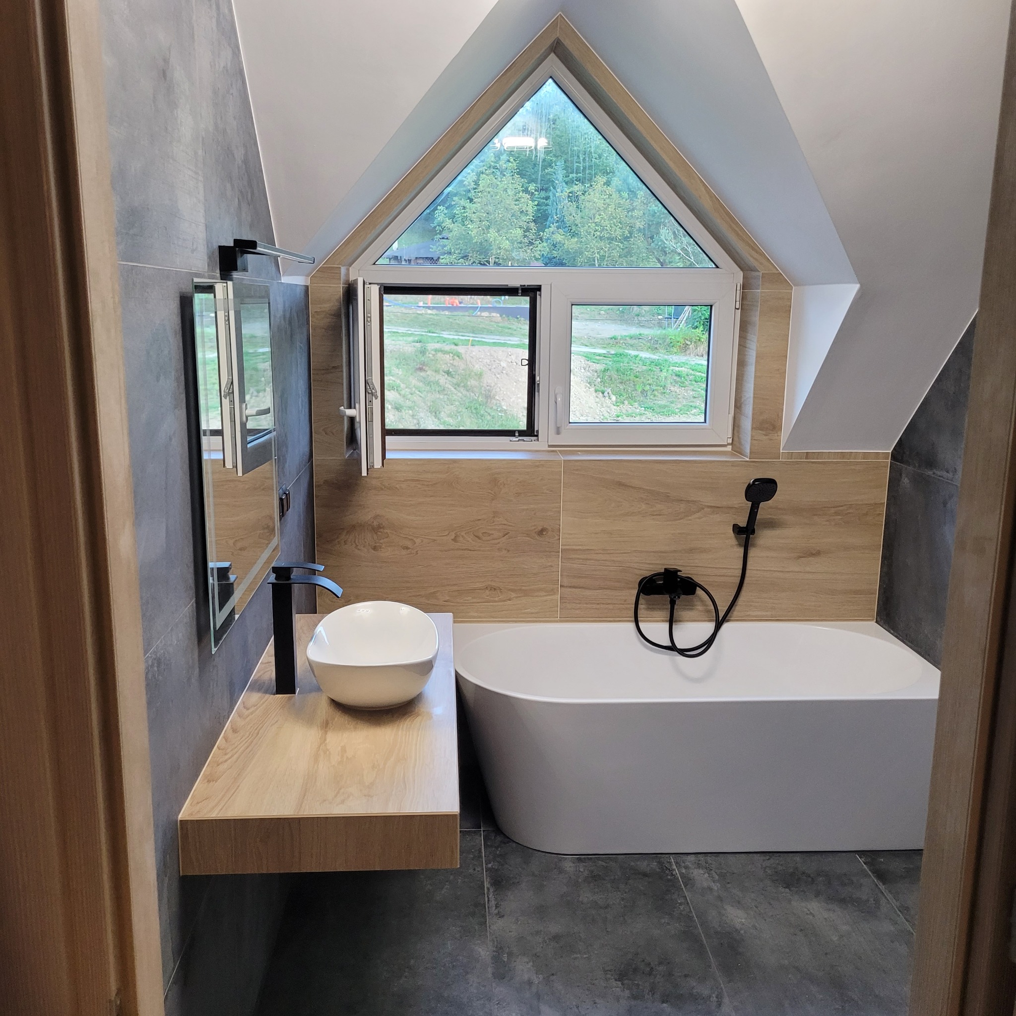 Nowoczesna łazienka na poddaszu z oknem trójkątnym, białą wanną i umywalką nablatową na drewnianym blacie. Szare i drewnopodobne płytki.