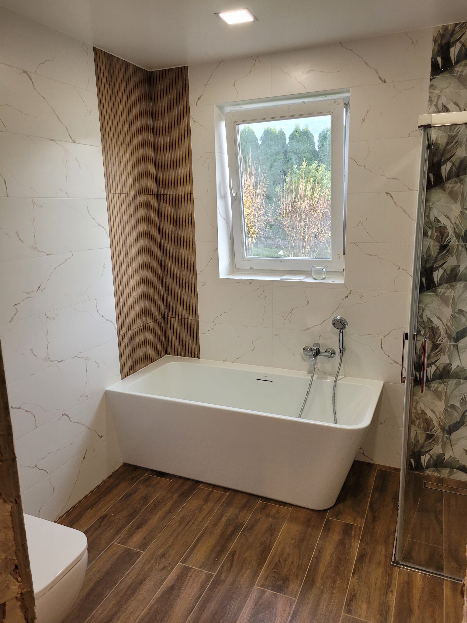 Nowoczesna łazienka z wolnostojącą wanną, drewnianą okładziną ścienną i marmurowymi płytkami. Widok na okno z zielenią. Drewnopodobna podłoga.