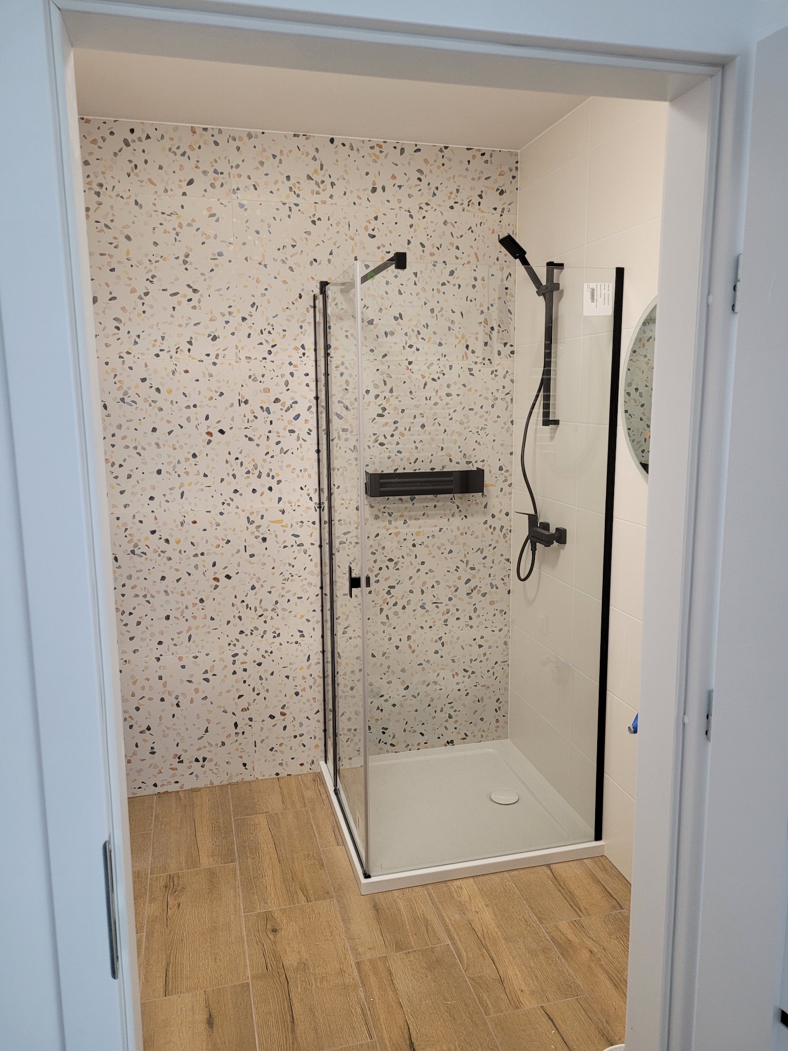 Nowoczesna kabina prysznicowa z czarną armaturą i deszczownicą, otoczona płytkami w stylu lastryko i drewnopodobną podłogą, widok zza otwartych drzwi.