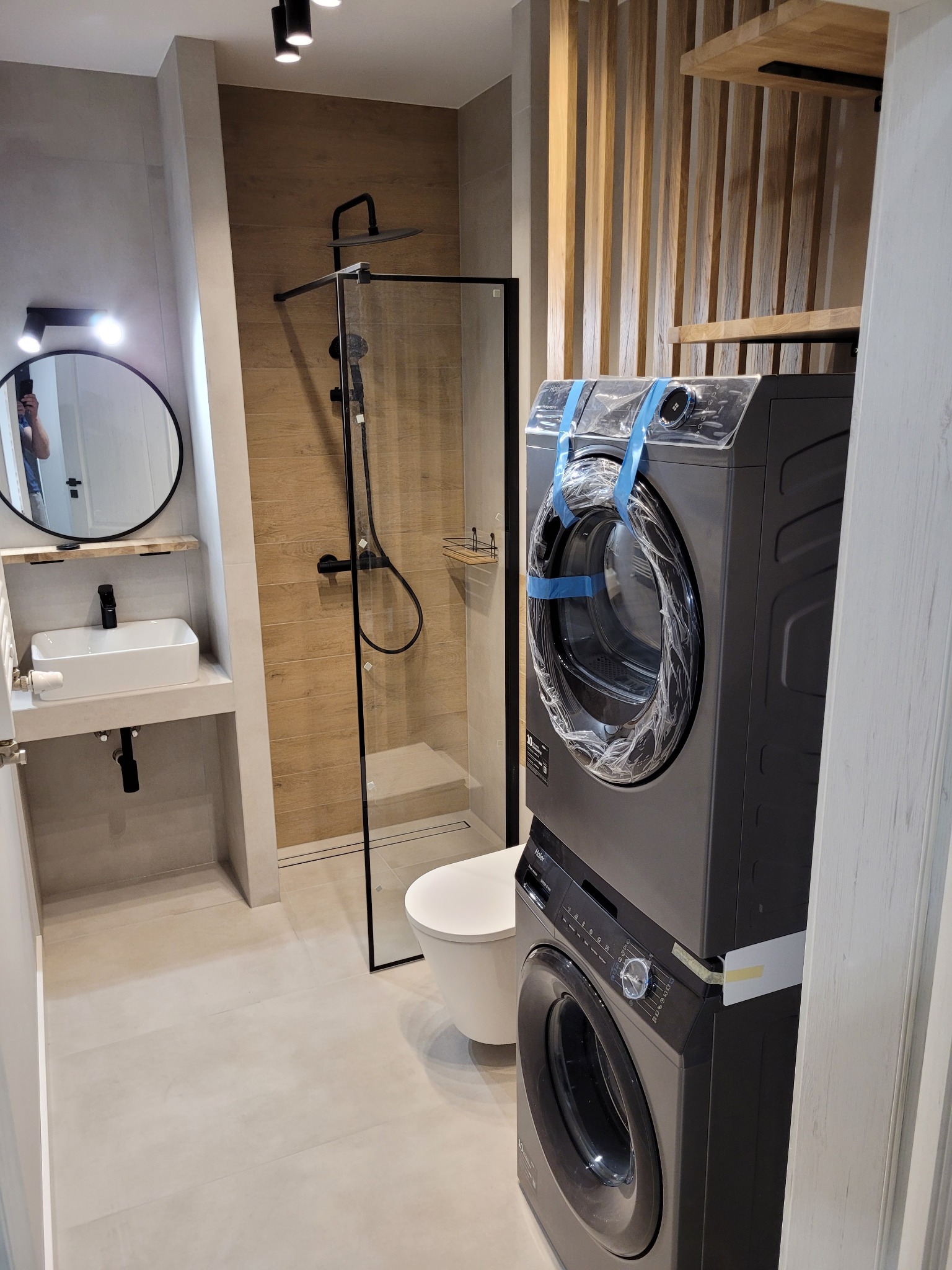 Wykończona łazienka z kabiną prysznicową typu walk-in, umywalką nablatową i toaletą. Na pierwszym planie pralka i suszarka w folii ochronnej.