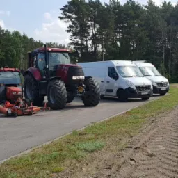 Czerwony traktor z kosiarką i dwa białe busy zaparkowane obok świeżo ułożonej trawy z rolki, w tle las.