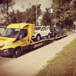 Żółta laweta marki IVECO z logo Procar przewozi trzy samochody osobowe: biały, czarny i czarny, na długiej platformie wzdłuż chodnika i ulicy w słoneczny dzień.