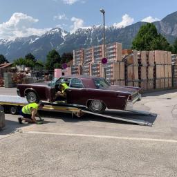 PROCAR STANISŁAW KICIŃSKI - Klasyczny, bordowy Lincoln Continental z otwartymi drzwiami, wciągany na niskopodwoziową lawetę w Turze, z pracownikami w kamizelkach odblaskowych, a w tle malowniczy widok na góry i palety z cegłami.