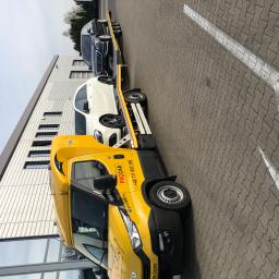 PROCAR STANISŁAW KICIŃSKI - Żółta laweta z dwoma samochodami (białym i czarnym) na platformie, zaparkowana przed nowoczesnym budynkiem. Widoczny branding firmy PROCAR.