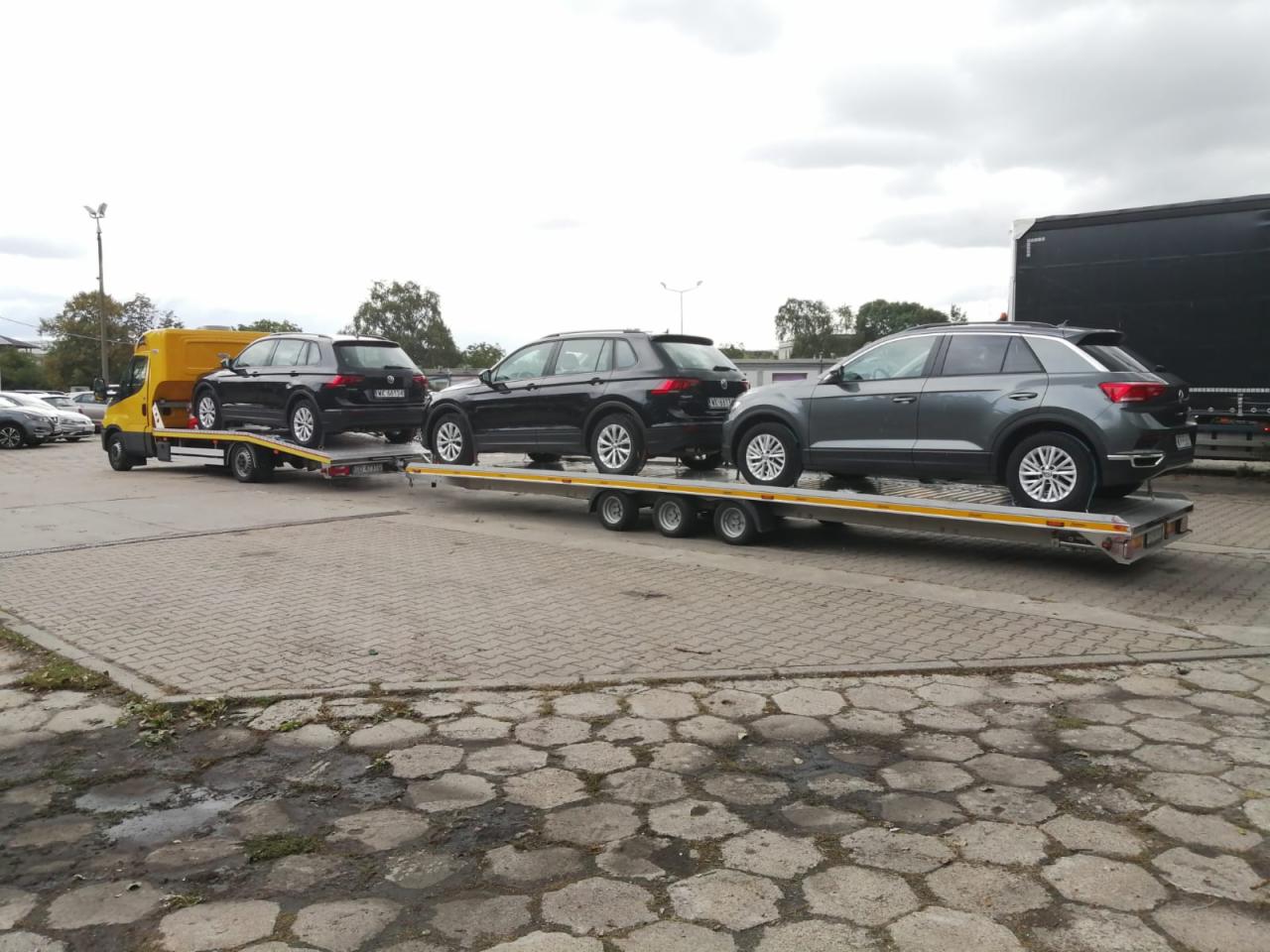 Żółta laweta z trzema samochodami osobowymi marki Volkswagen, zaparkowana na betonowym placu w pochmurny dzień.