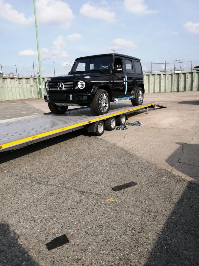 Czarny Mercedes G-klasa zabezpieczony niebieskimi pasami na żółtej platformie lawety, widok z boku i dołu, w tle betonowy plac i błękitne niebo z chmurami.
