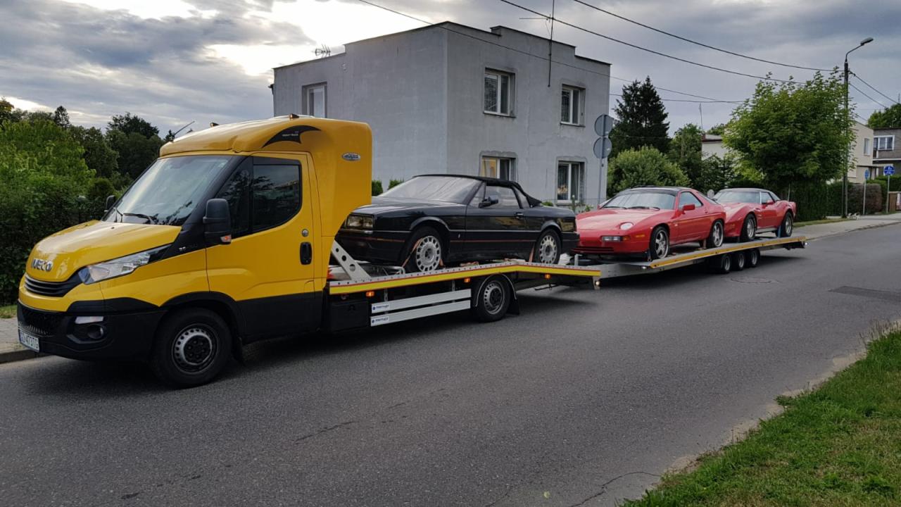 Żółta ciężarówka pomocy drogowej Iveco przewozi na platformie trzy sportowe samochody: czarny Cadillac Allante i dwa czerwone Porsche 928 na tle szarego budynku mieszkalnego.