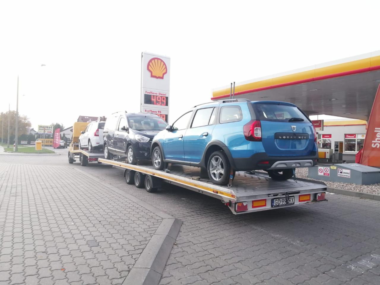Laweta z trzema samochodami Dacia, w tym niebieski Logan MCV, na tle stacji benzynowej Shell.