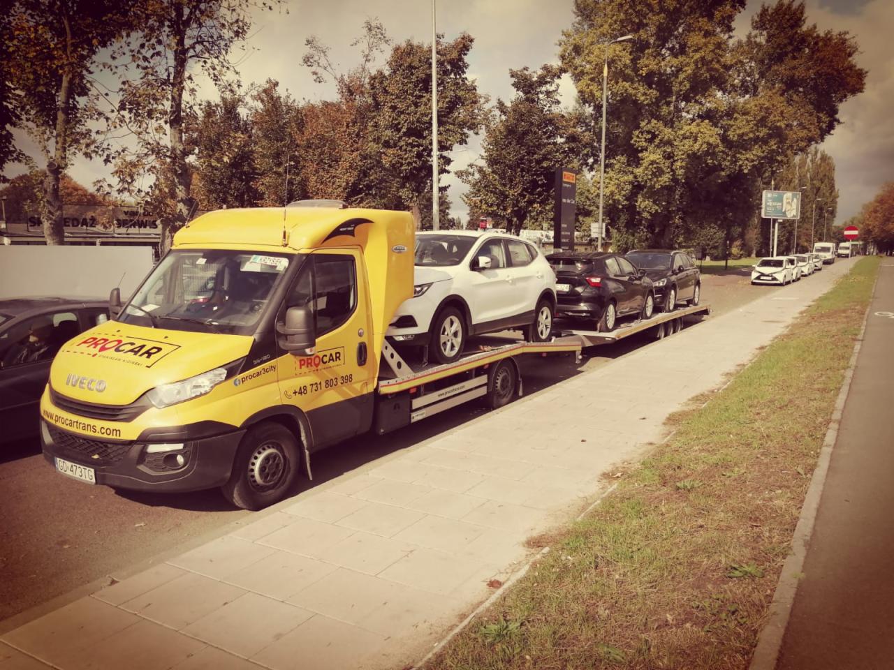 Żółta laweta marki IVECO z logo Procar przewozi trzy samochody osobowe: biały, czarny i czarny, na długiej platformie wzdłuż chodnika i ulicy w słoneczny dzień.