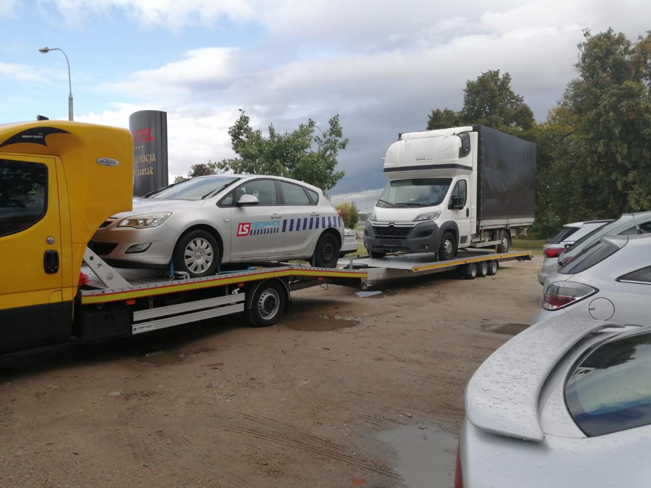 Żółta laweta z srebrnym samochodem osobowym marki Opel i biały samochód dostawczy na platformie, widok z boku, pochmurny dzień.