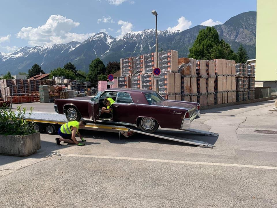 Klasyczny, bordowy Lincoln Continental z otwartymi drzwiami, wciągany na niskopodwoziową lawetę w Turze, z pracownikami w kamizelkach odblaskowych, a w tle malowniczy widok na góry i palety z cegłami.