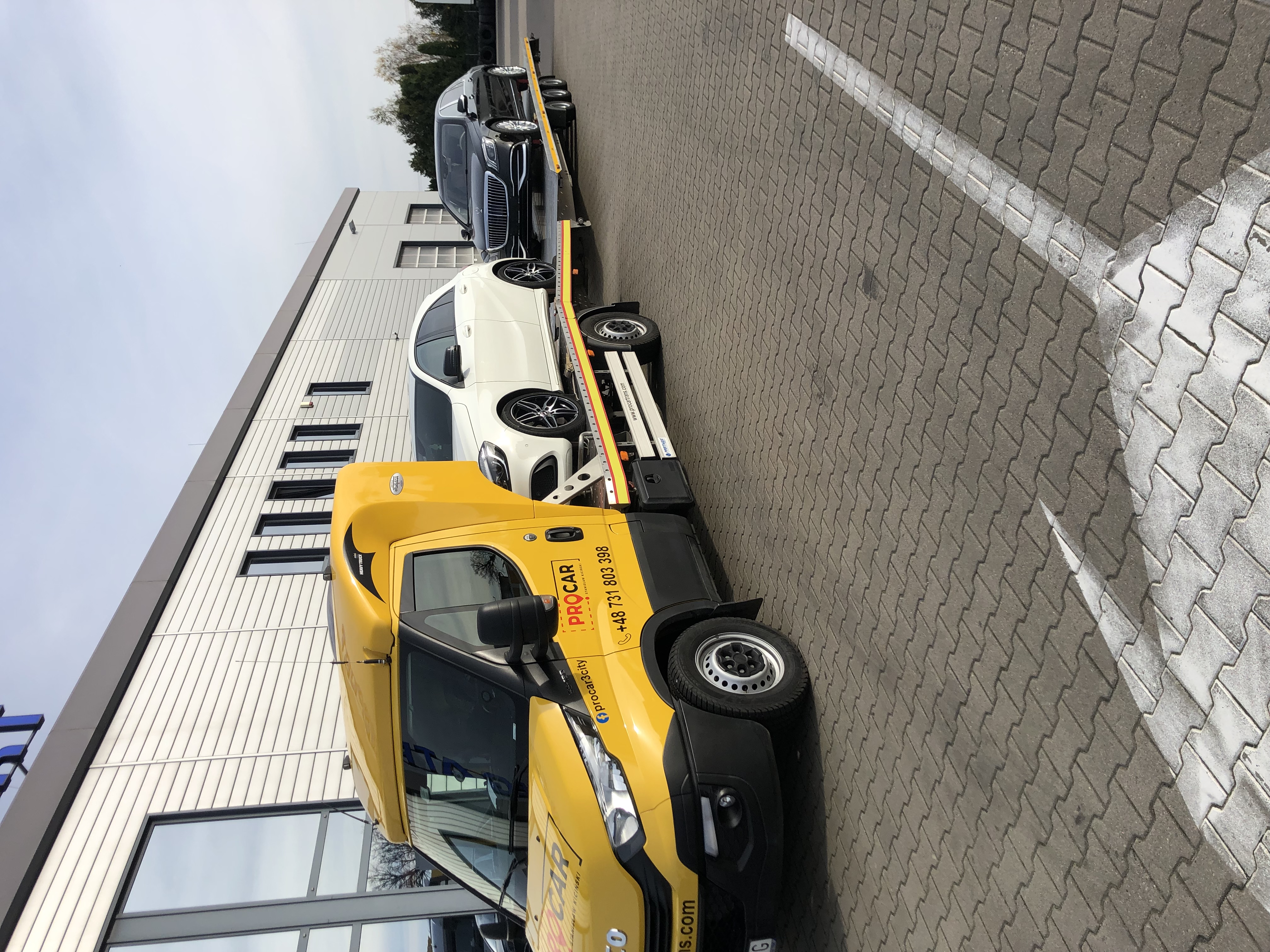 Żółta laweta z dwoma samochodami (białym i czarnym) na platformie, zaparkowana przed nowoczesnym budynkiem. Widoczny branding firmy PROCAR.