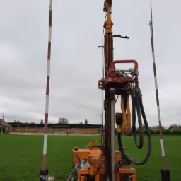 Pomarańczowa wiertnica do studni głębinowych na trawiastym terenie z widocznymi tyczkami wyznaczającymi obszar robót, stadion w tle pod pochmurnym niebem.