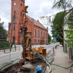 Żółta maszyna wiertnicza na gąsienicach pracuje przy wykopie wypełnionym wodą w Zawoni, na tle ceglanego kościoła z wieżą zegarową.