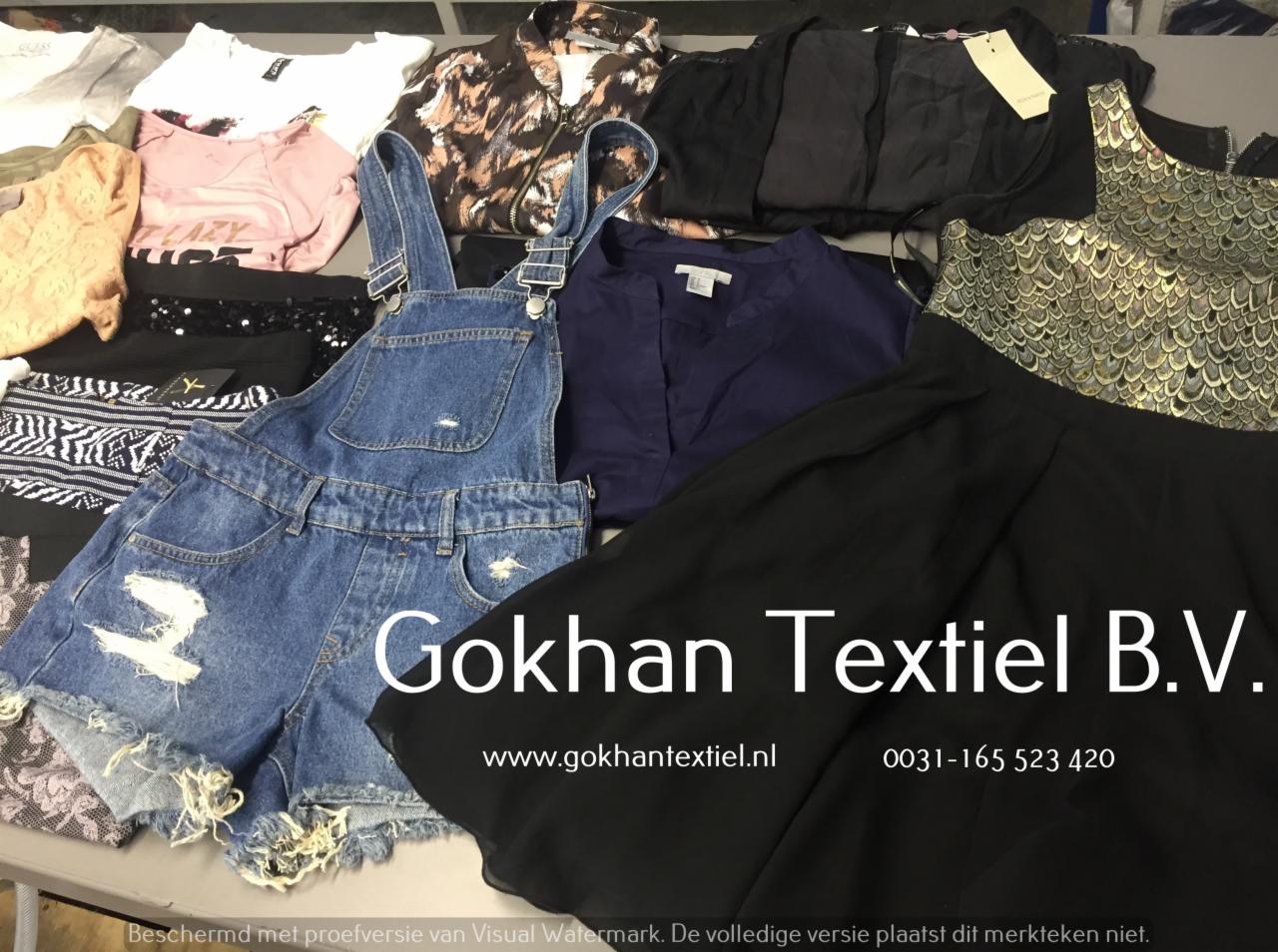 Stos ubrań, w tym dżinsowe ogrodniczki z przetarciami, koszulka z napisem 'Lazy' i czarna spódnica, ułożone na stole z widocznym logo i danymi kontaktowymi firmy Gokhan Textiel B.V.