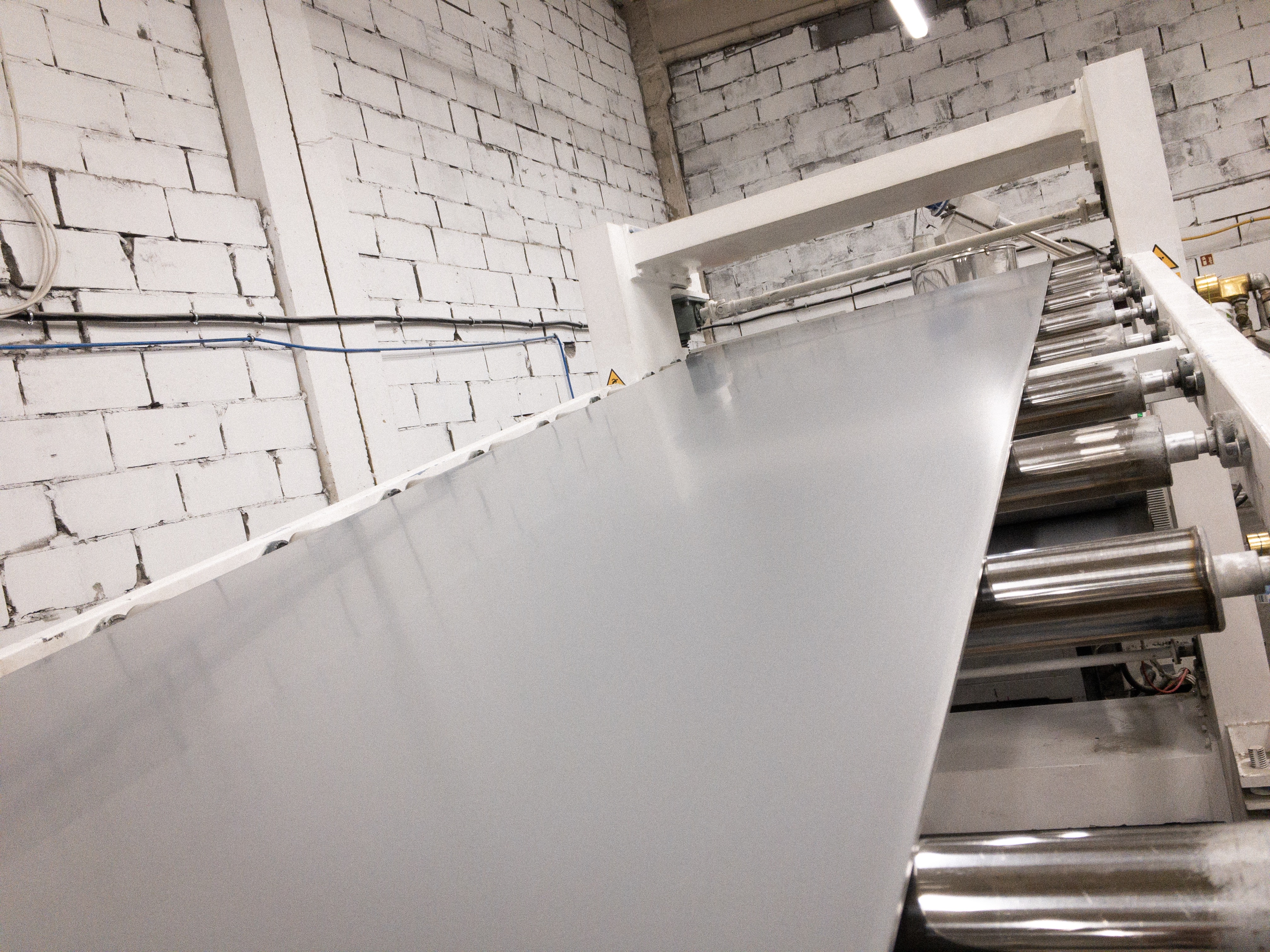 Srebrna folia przechodząca przez system rolek w maszynie produkcyjnej, widoczna ceglana ściana w tle.