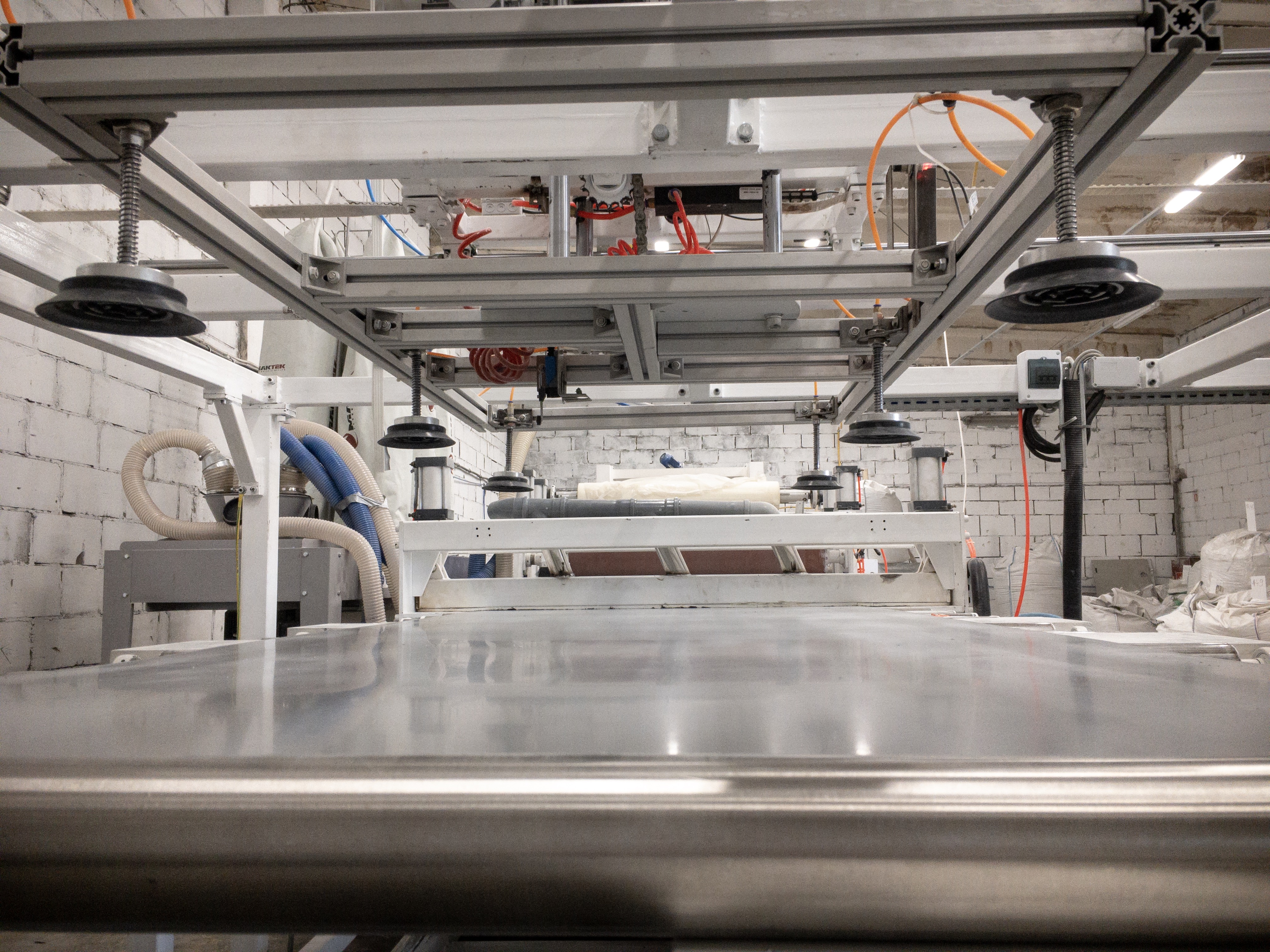 Widok z dołu na linię produkcyjną z metalową konstrukcją, przyssawkami i rurami, w tle białe worki z materiałem.