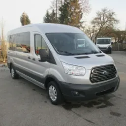 Srebrny, fabrycznie nowy Ford Transit z długim rozstawem osi, zaparkowany na betonowym placu w słoneczny dzień, z innymi pojazdami dostawczymi w tle.