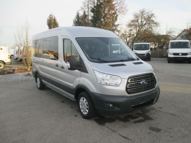 Srebrny, fabrycznie nowy Ford Transit z długim rozstawem osi, zaparkowany na betonowym placu w słoneczny dzień, z innymi pojazdami dostawczymi w tle.