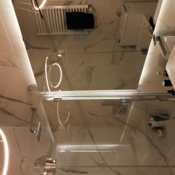 Remonty Tadeusz Nowak - Nowoczesna łazienka z marmurowymi płytkami, szklanym panelem prysznicowym z podświetleniem LED, białym grzejnikiem drabinkowym z czarnym ręcznikiem i minimalistyczną umywalką z czarną armaturą.