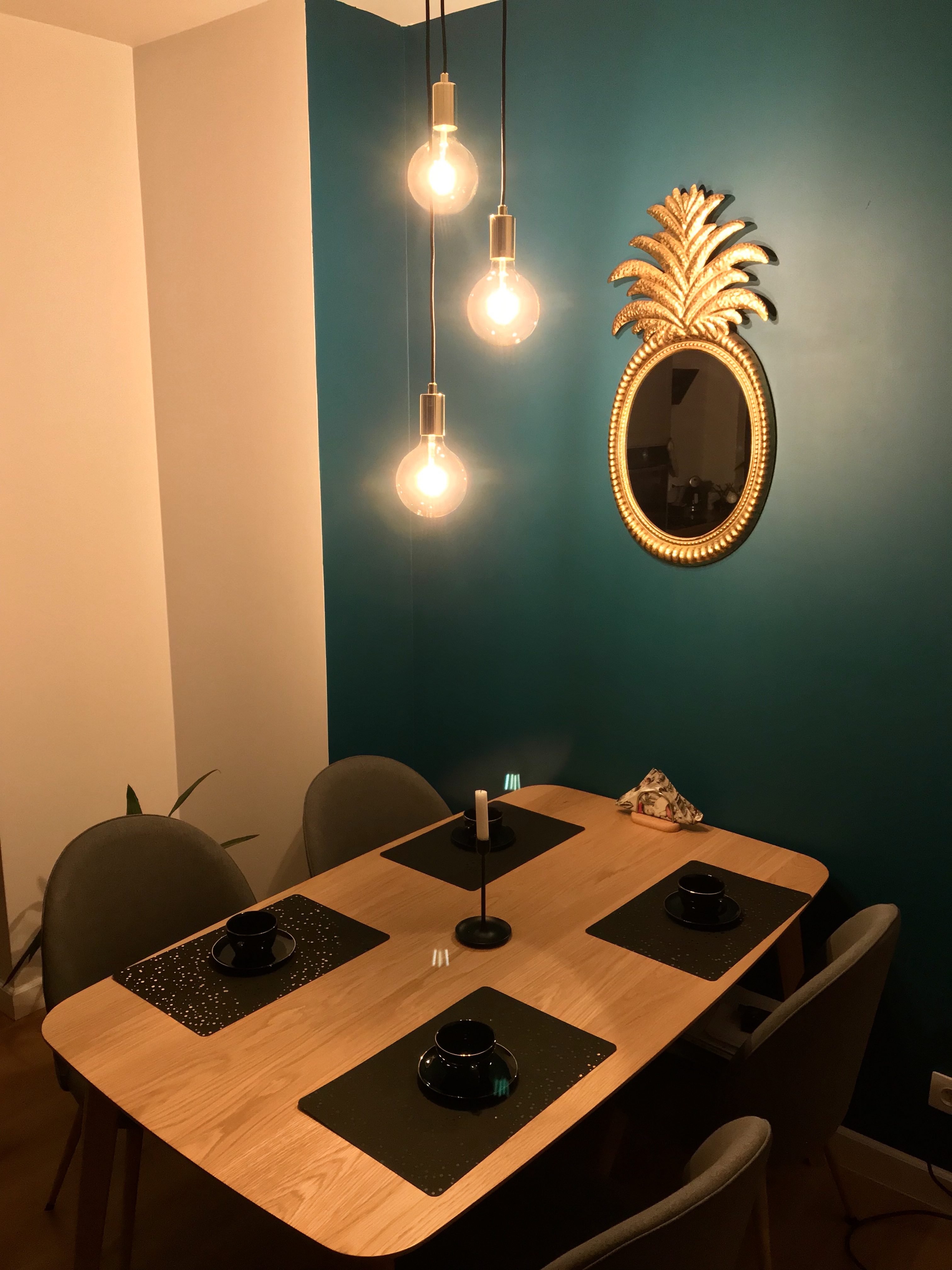 Jadalnia z drewnianym stołem nakrytym dla czterech osób, czarne podkładki z białymi kropkami, czarne filiżanki, świecznik z białą świecą, ściana w kolorze turkusowym z lustrem w złotej ramie...