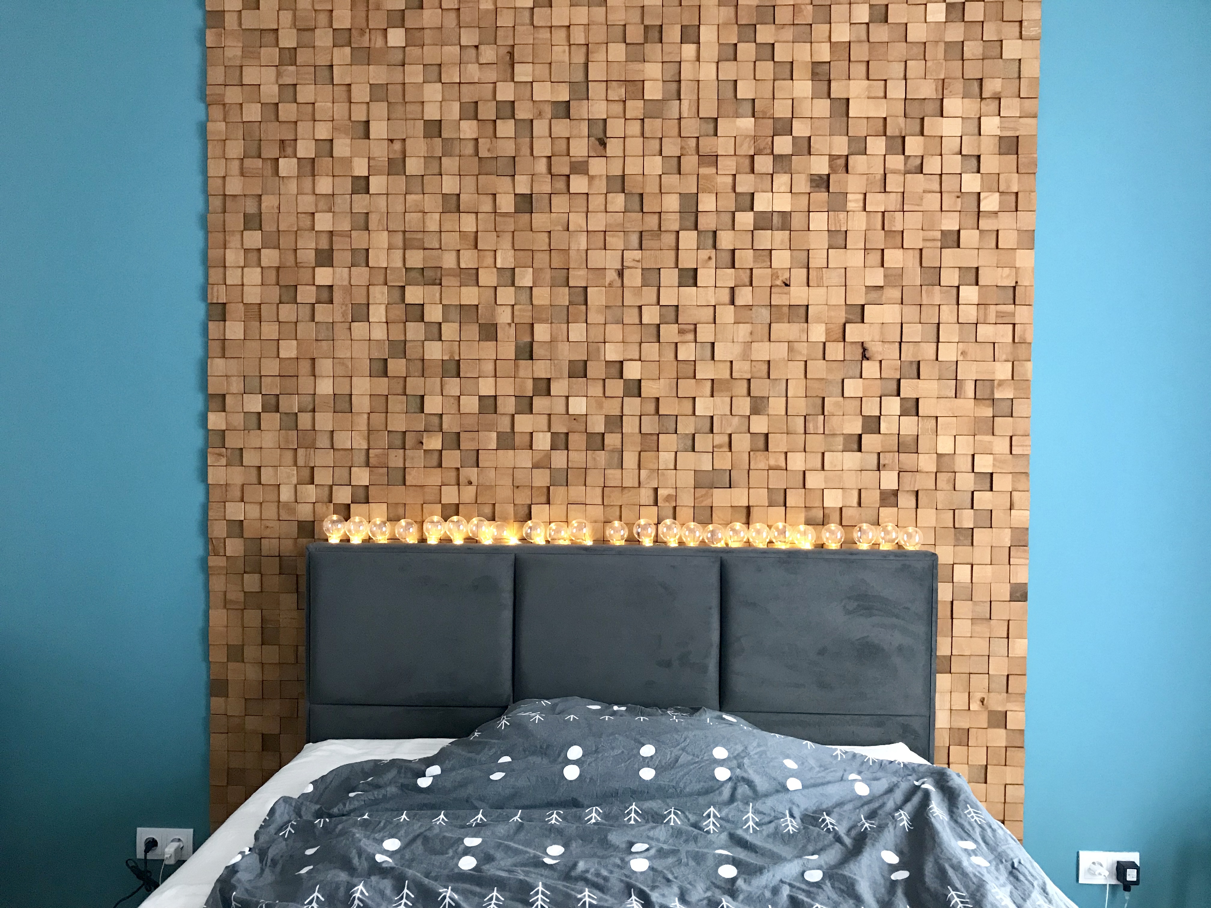 Sypialnia z dekoracyjną ścianą z drewnianych kostek, oświetlona girlandą żarówkową nad wezgłowiem łóżka, na tle turkusowej ściany.
