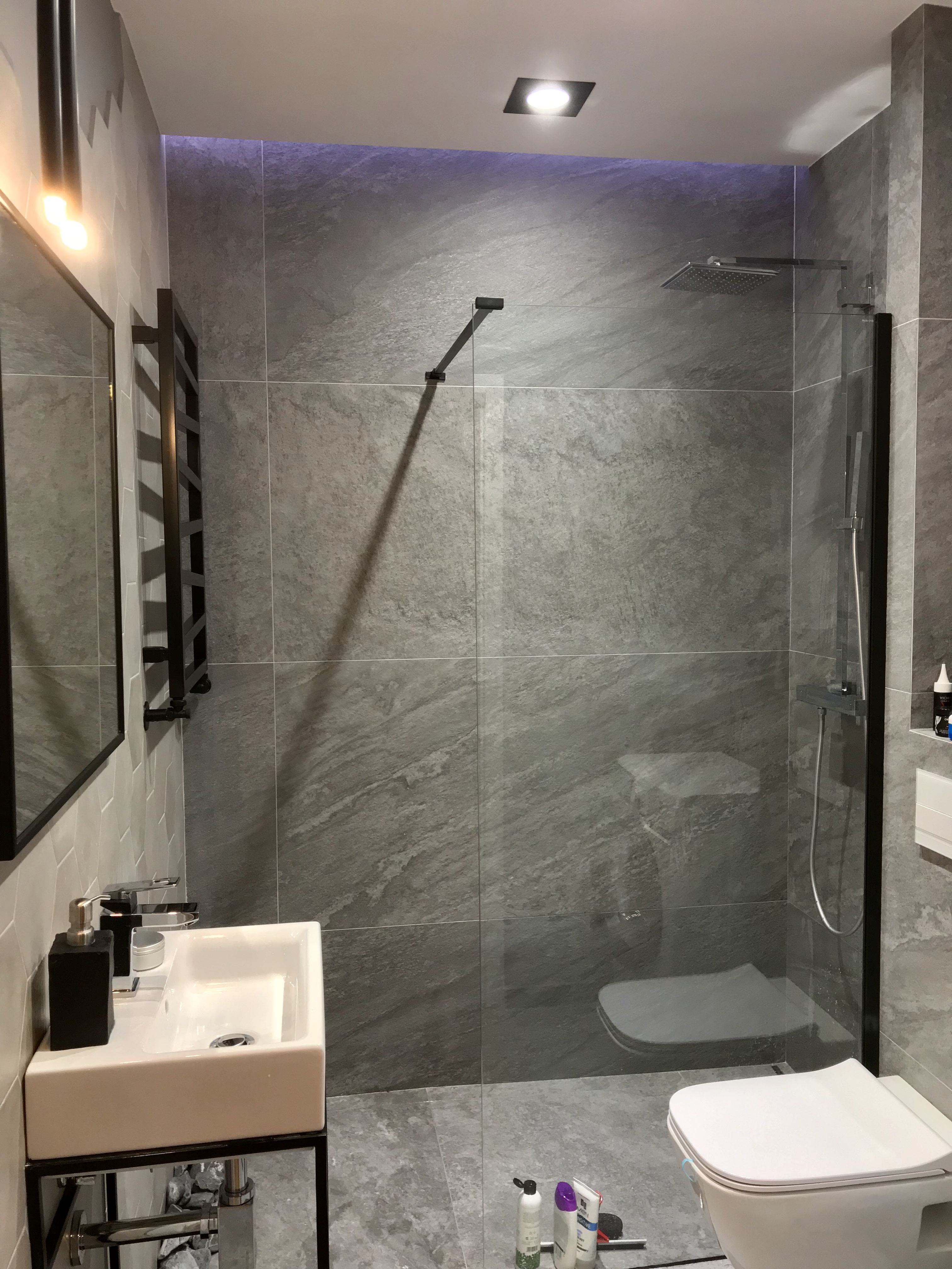 Nowoczesna łazienka z szarymi płytkami imitującymi kamień, kabiną prysznicową typu walk-in z czarnymi elementami, białą umywalką na czarnym stelażu i grzejnikiem drabinkowym w tym samym kolorze...