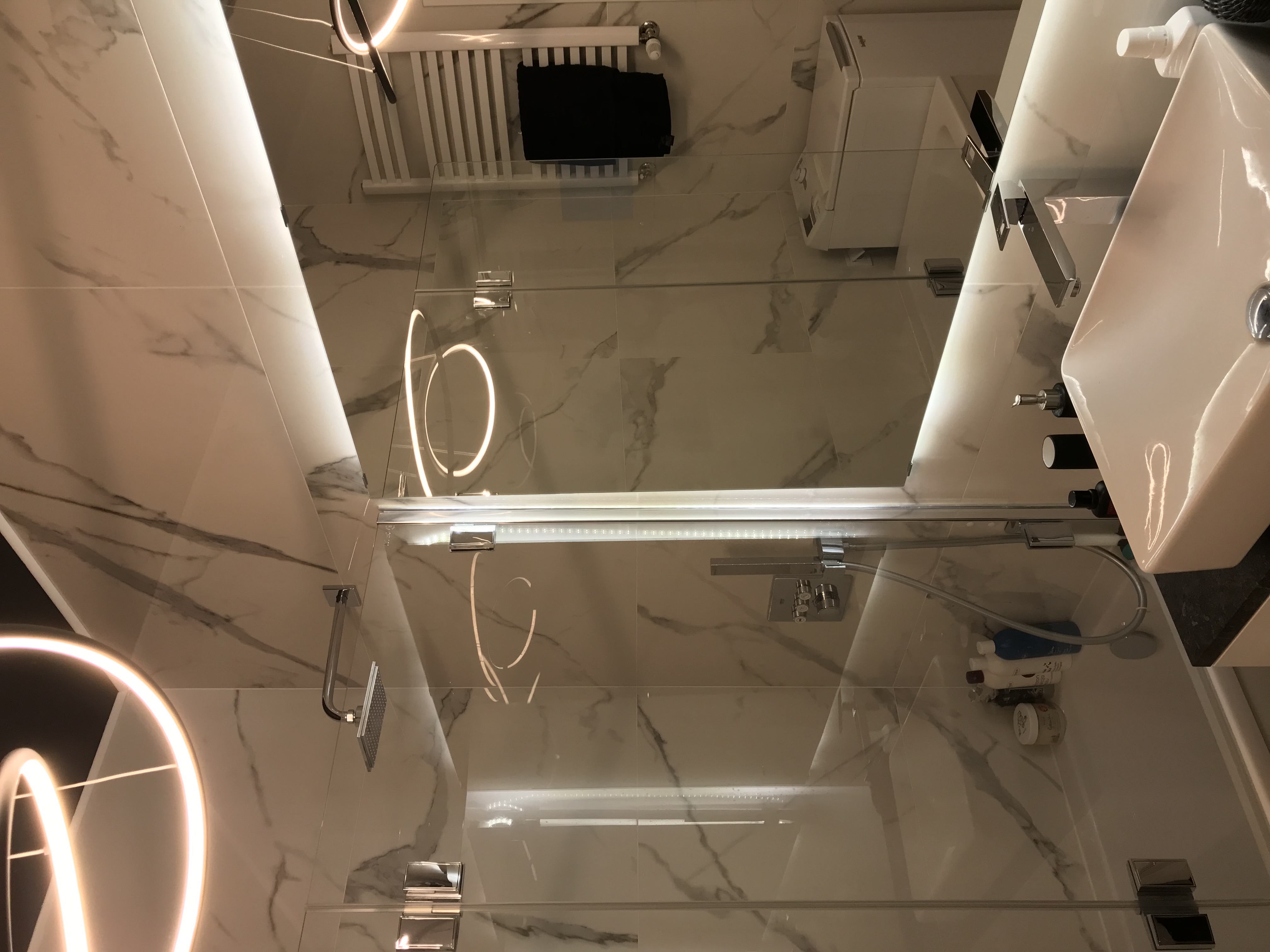 Nowoczesna łazienka z marmurowymi płytkami, szklanym panelem prysznicowym z podświetleniem LED, białym grzejnikiem drabinkowym z czarnym ręcznikiem i minimalistyczną umywalką z czarną armaturą.