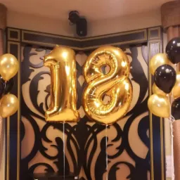 Dekoracja na 18 urodziny: Złote balony foliowe w kształcie cyfr '1' i '8' otoczone bukietami złotych i czarnych balonów lateksowych na tle dekoracyjnej ściany z ornamentem.