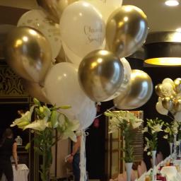 Odlotowy balon - Dekoracja stołu na uroczystość w Warszawie: białe lilie w smukłych wazonach i balony w kolorach bieli i złota, niektóre z napisem, ustawione na długim stole z białym obrusem i różowymi krzesłami.