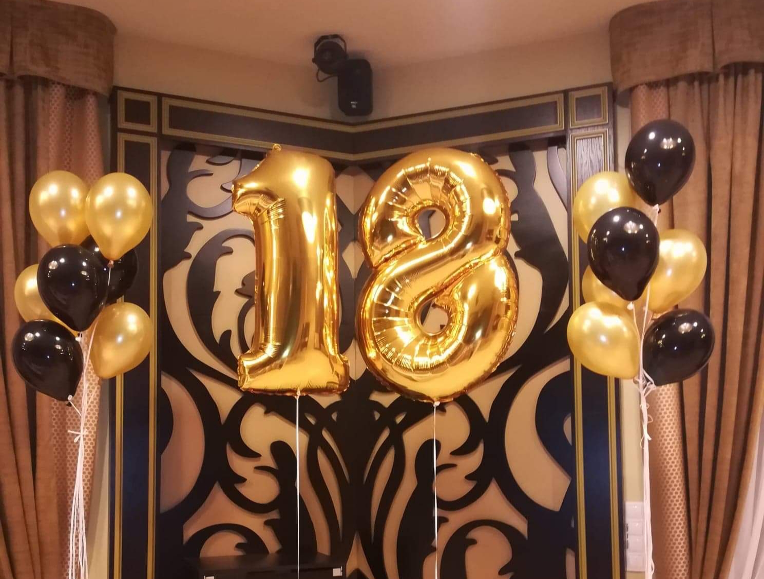 Dekoracja na 18 urodziny: Złote balony foliowe w kształcie cyfr '1' i '8' otoczone bukietami złotych i czarnych balonów lateksowych na tle dekoracyjnej ściany z ornamentem.