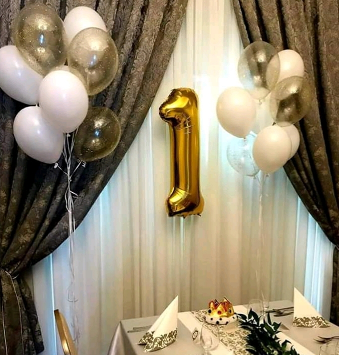 Dekoracja stołu na pierwsze urodziny z białymi i złotymi balonami oraz złotą cyfrą jeden, ustawiona na tle drapowanych zasłon.