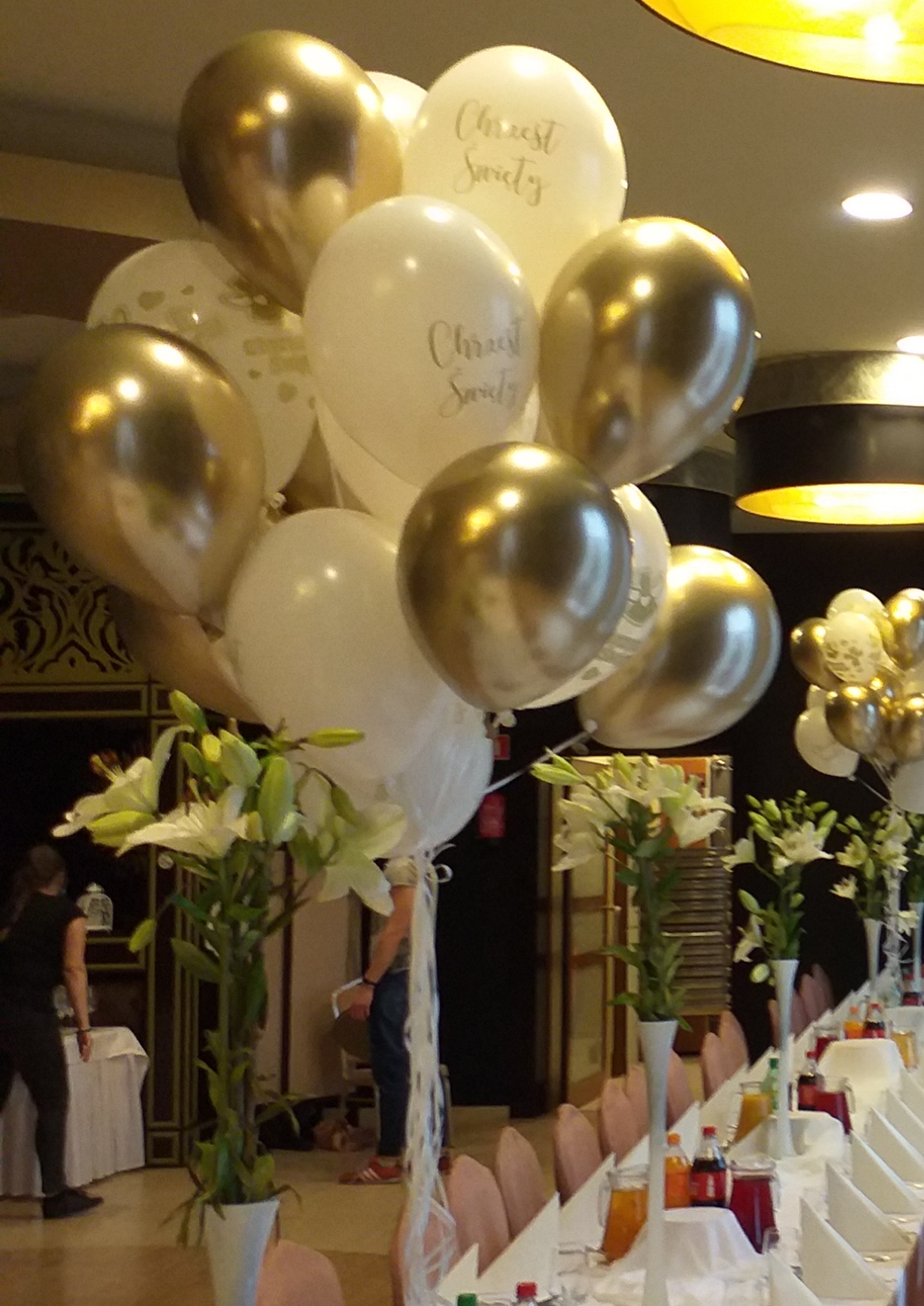 Dekoracja stołu na uroczystość w Warszawie: białe lilie w smukłych wazonach i balony w kolorach bieli i złota, niektóre z napisem, ustawione na długim stole z białym obrusem i różowymi krzesłami.