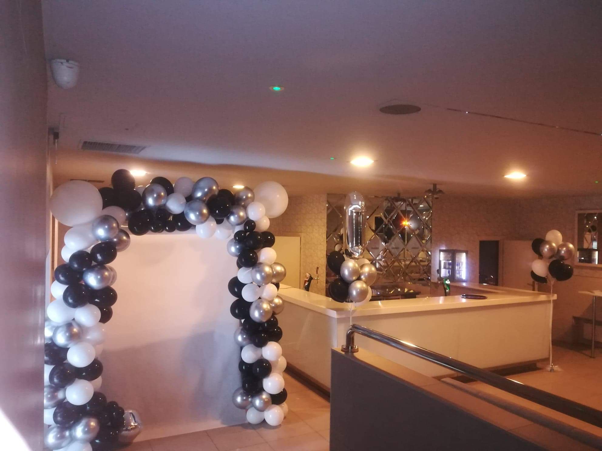 Dekoracja balonowa w odcieniach czerni, bieli i srebra tworząca łuk wejściowy, z dodatkowymi balonami przy barze z lustrzaną ścianą w tle.
