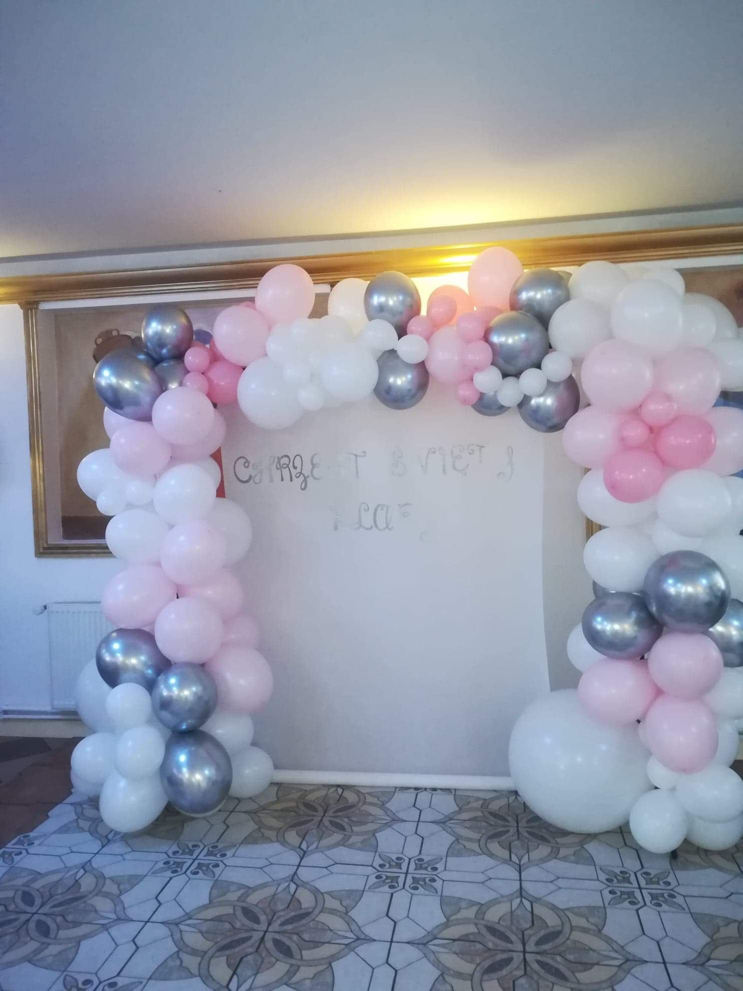Dekoracja balonowa w kształcie łuku z białych, różowych i srebrnych balonów na tle białej ściany z napisem, widoczna na tle wzorzysta podłoga.