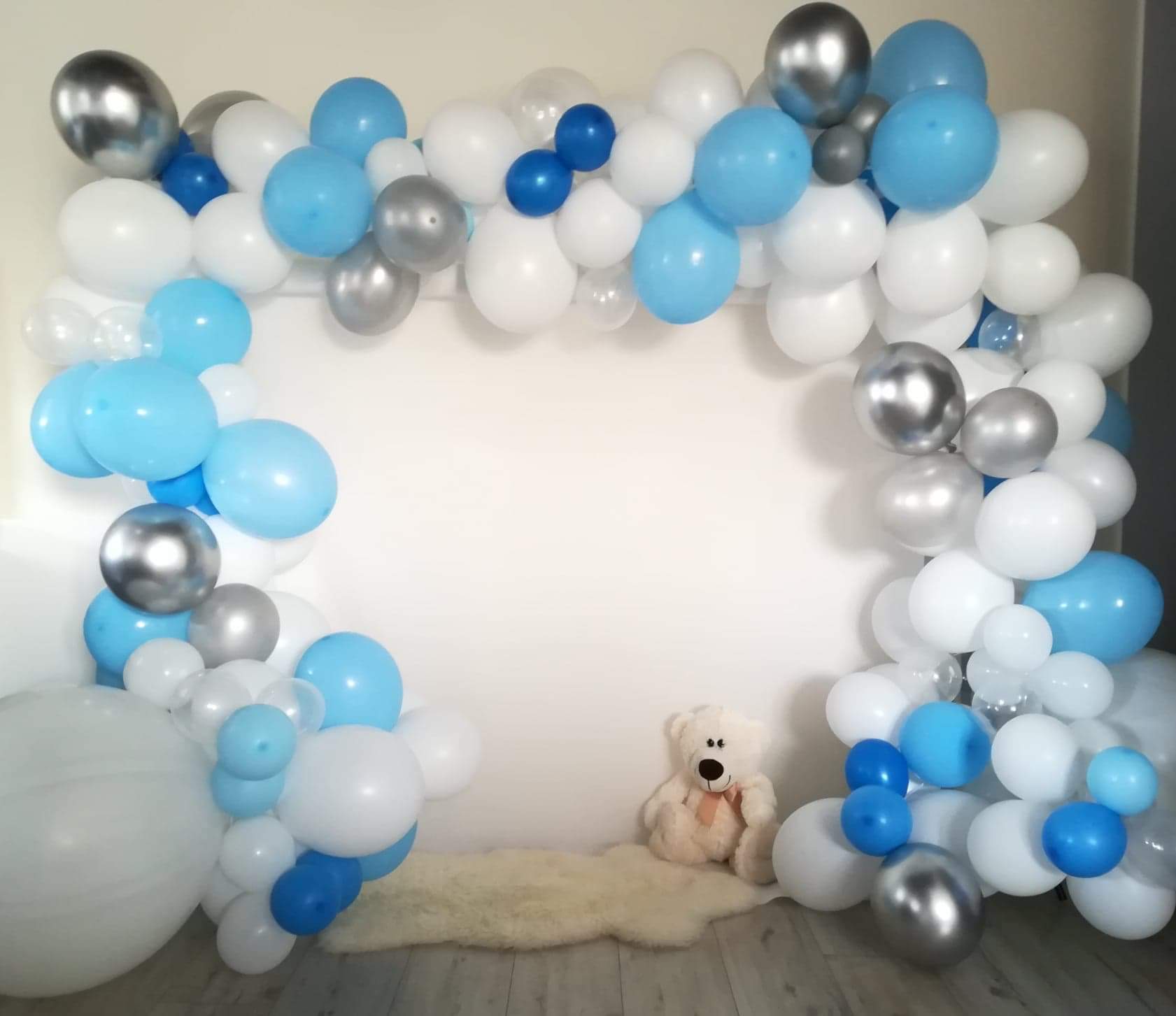 Dekoracja z białych, błękitnych i srebrnych balonów ułożonych w łuk, z pluszowym misiem siedzącym na białym dywaniku w tle, na tle białej ściany.